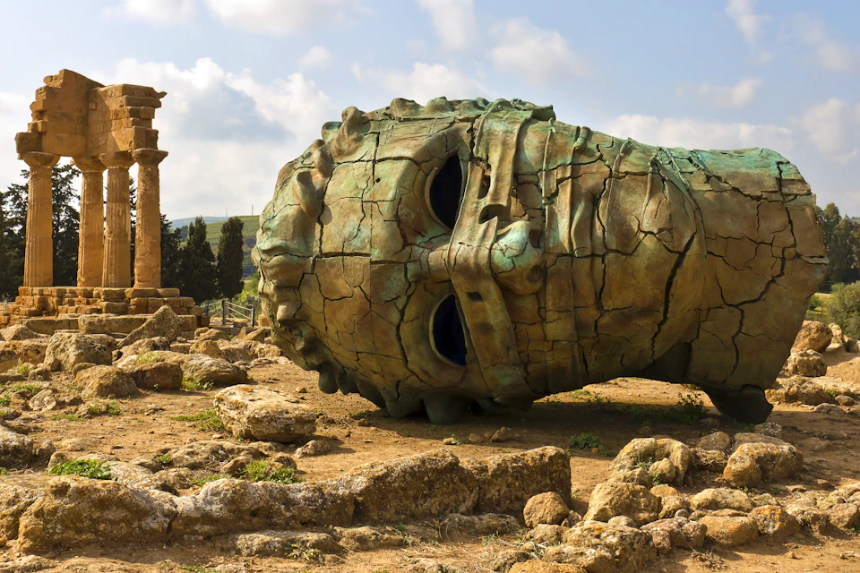 A large, cracked bronze statue head lies on the ground among ancient ruins under a partly cloudy sky.