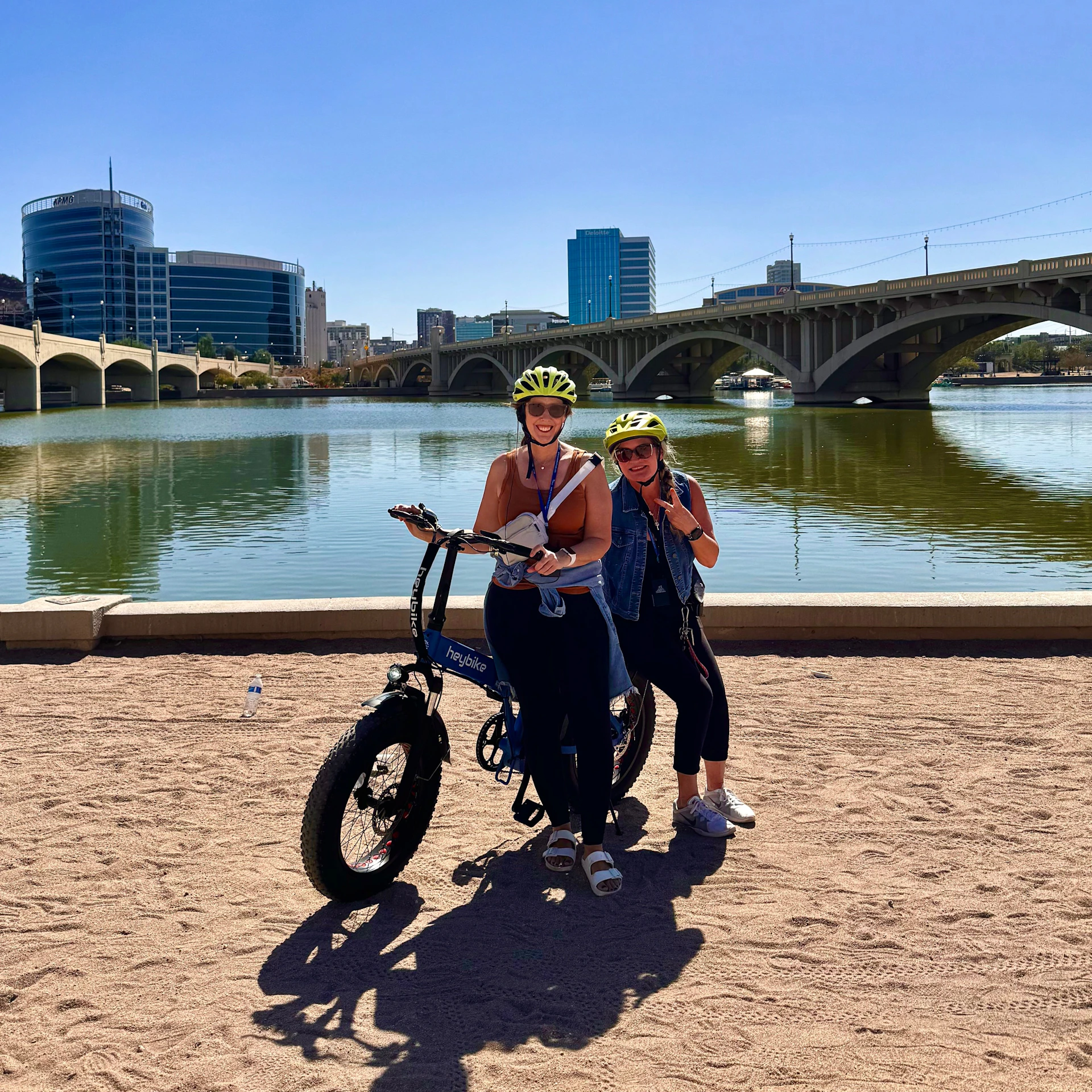 Two people wearing helmets stand by an electric bike on a sandy path near a river, with a bridge and buildings in the background.