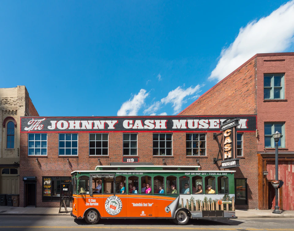 A green and orange trolley bus with tourists passing in front of the Johnny Cash Museum, a brick building with a clear sign.