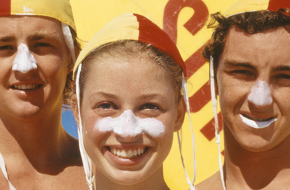 Three people wearing red and yellow swim caps and zinc on their noses, standing in front of a yellow surfboard.