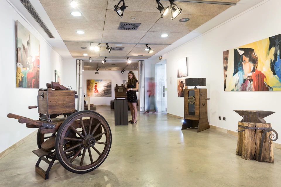 A woman in a black dress explores an art gallery with various sculptures and paintings on white walls, under track lighting.