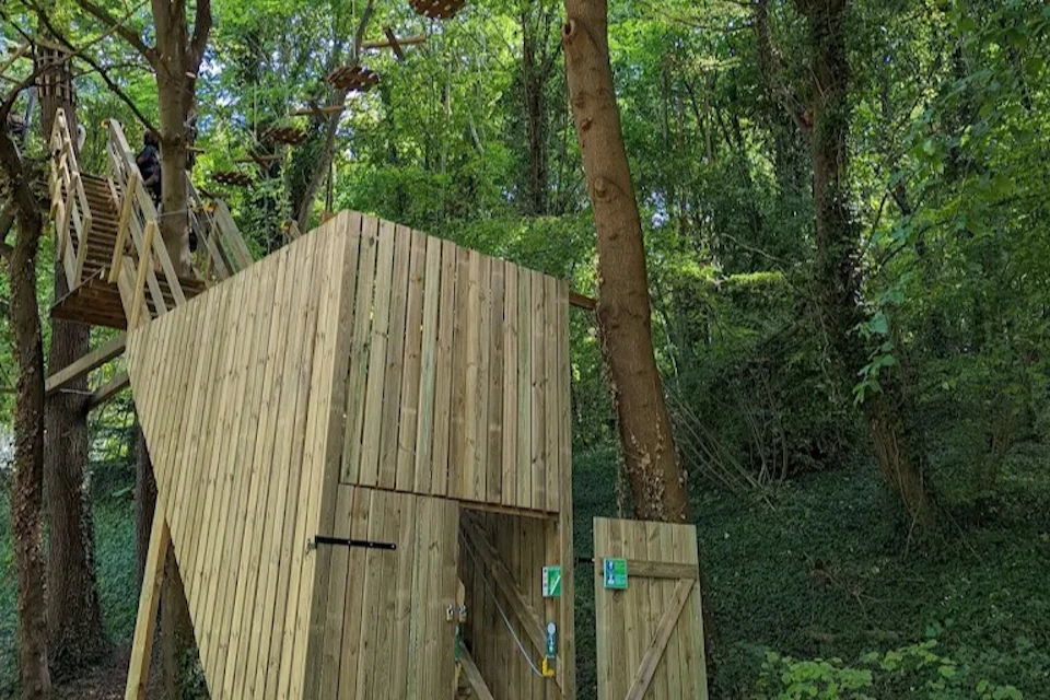 Wooden structures and platforms in a forest, with a rope course between trees. Dense greenery surrounds the area.
