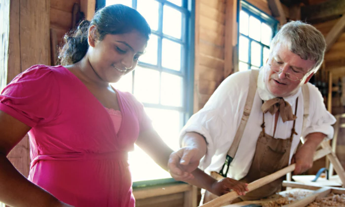 An older man is teaching woodworking to a child inside a well-lit workshop with wooden walls and large windows.