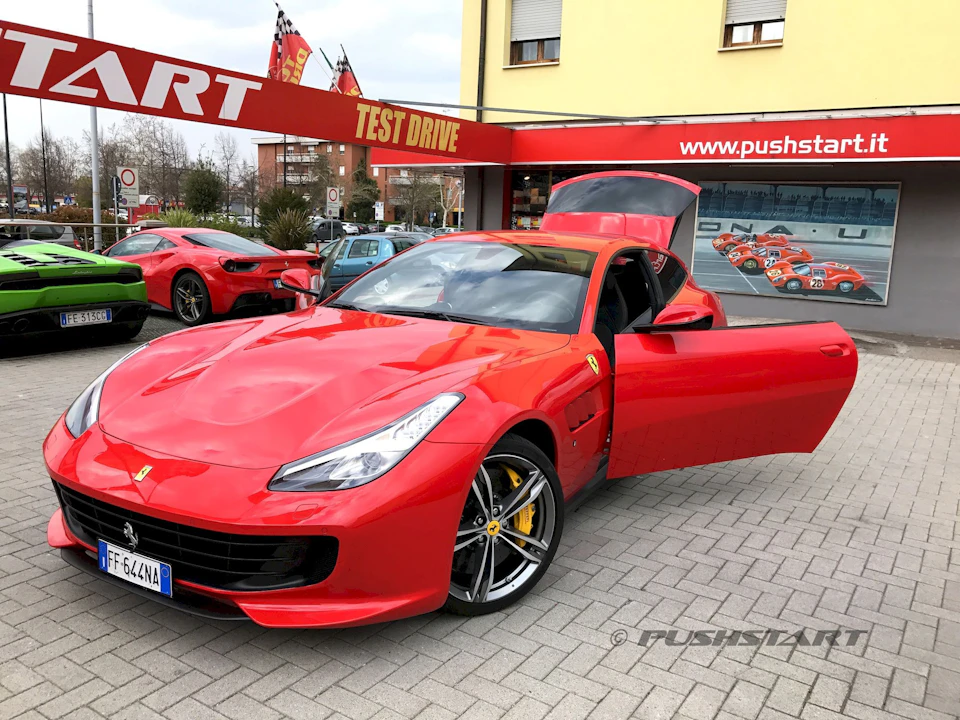 Red Ferrari with open doors and a raised rear hatch, parked in front of a test drive center.