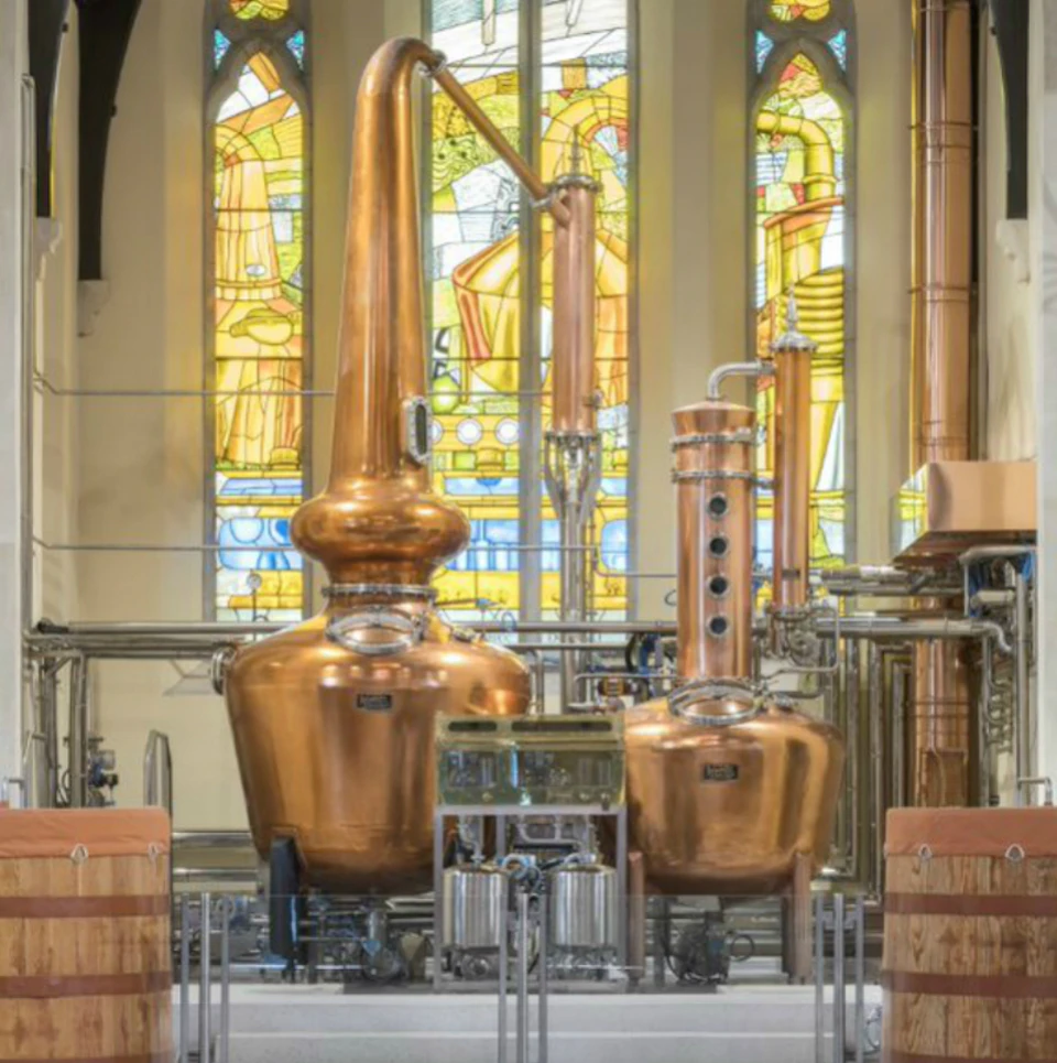 Two large copper distillation stills in front of stained glass windows inside a distillery or brewery.