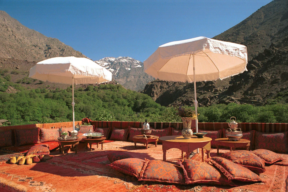 Outdoor seating area with ornate cushions, low tables, and white umbrellas, set against a mountainous backdrop with clear blue sky.