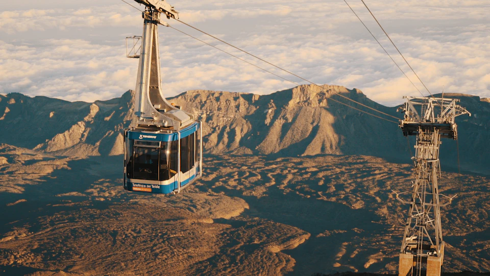 A blue cable car labeled "Teleférico del Teide" suspended over a rugged mountain landscape beneath a cloudy sky.