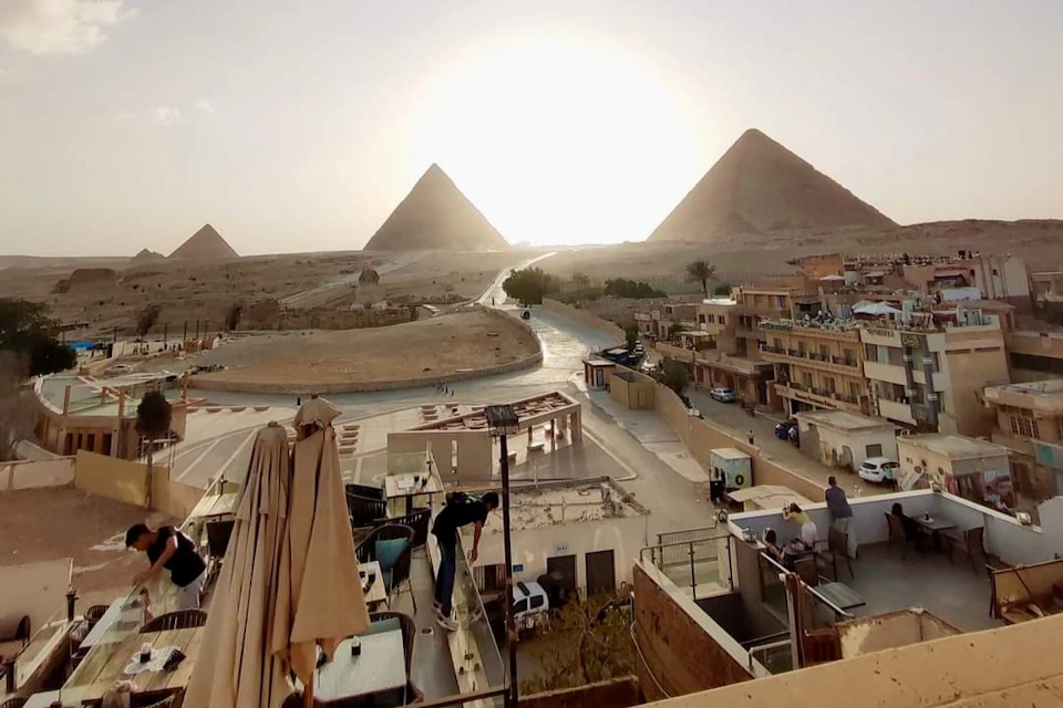 Three pyramids under a setting sun with a road leading toward them, flanked by buildings and terraces with people.