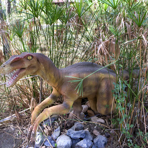 En dinosauriemodell med öppen mun ligger bland höga gräs och stenar i ett skogsområde.