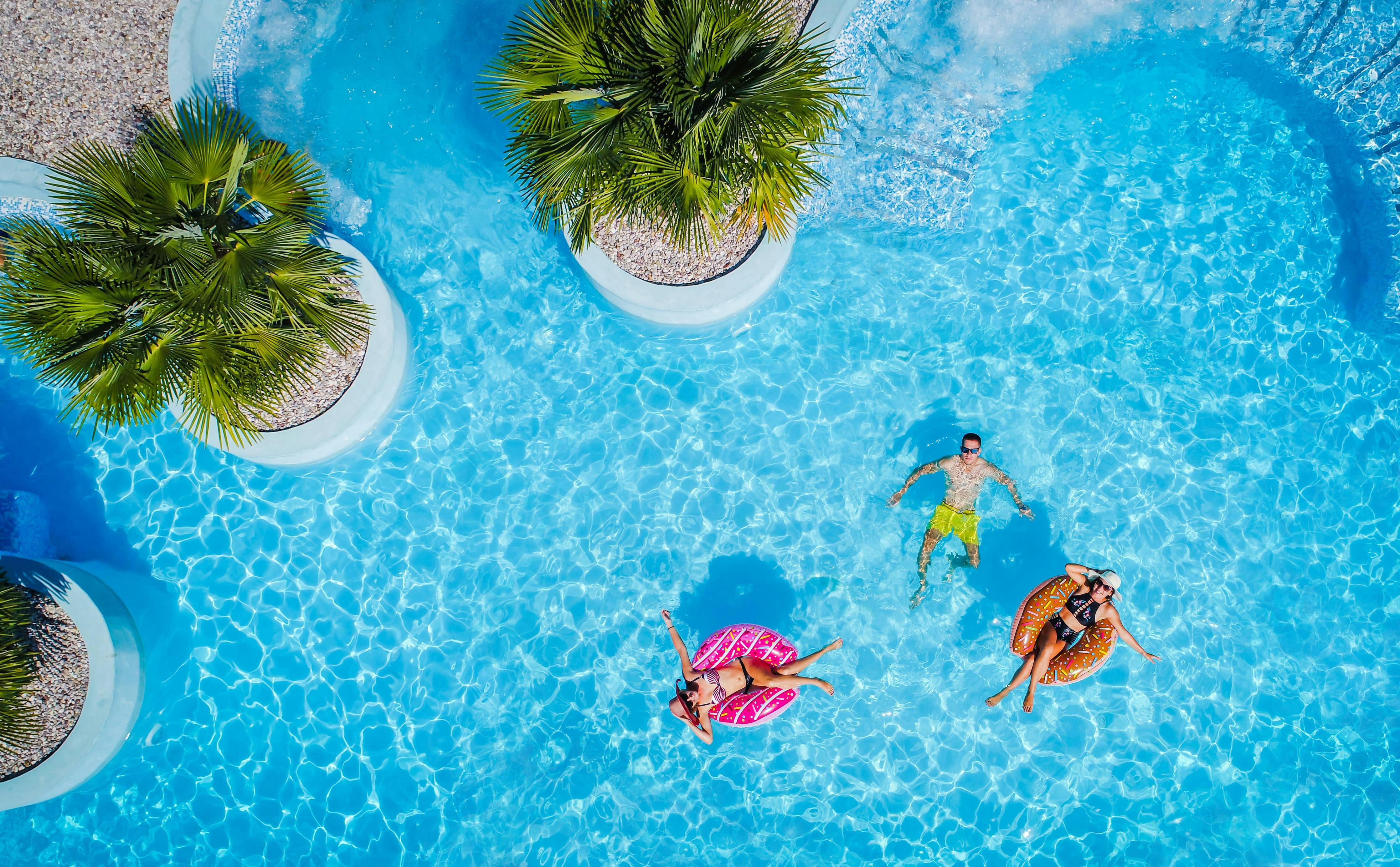 Drei Personen entspannen sich in einem Pool, zwei davon auf Schlauchbooten und eine schwimmend. Palmen säumen den Rand. Das Wasser kräuselt sich um sie herum.