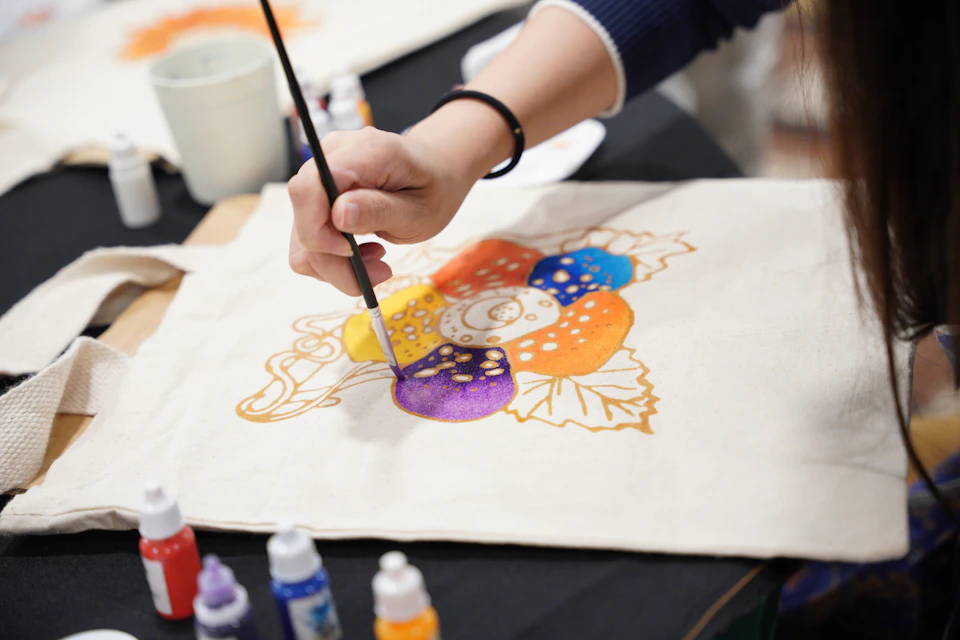 A person painting a colorful design on fabric with a brush, surrounded by paint bottles.
