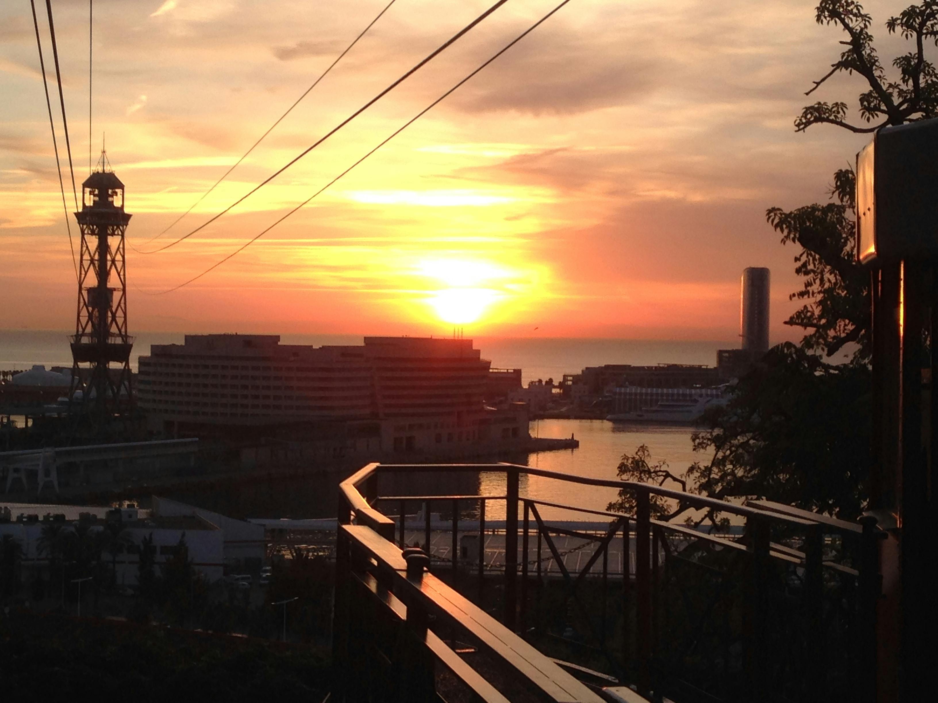 Západ slunce nad nábřežním panoramatem města s budovami, vodou, zábradlím v popředí a elektrickým vedením nahoře.