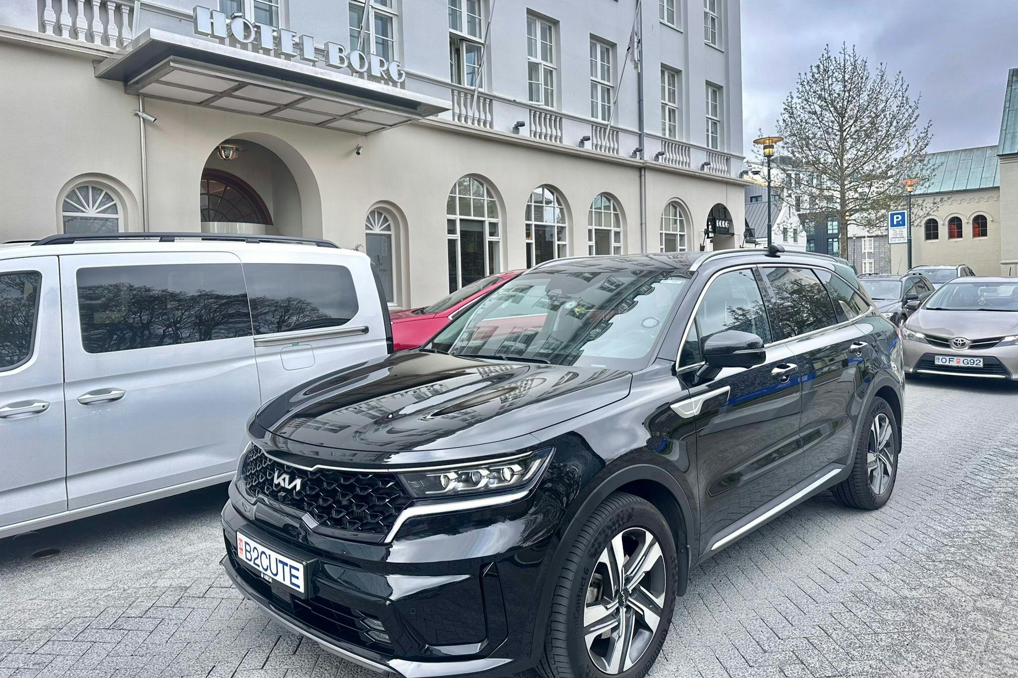 A black Kia SUV with a custom "B2CUTE" license plate is parked in front of the Hotel Borg. Nearby are a white van and a red car.