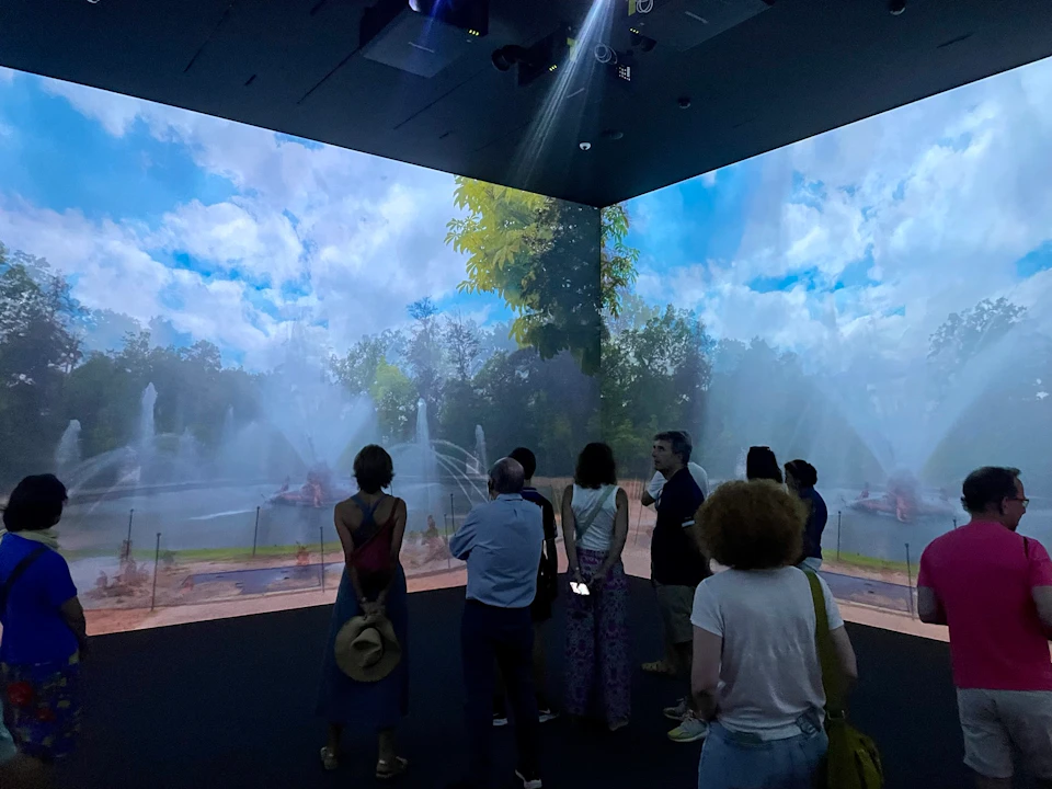 A group of people viewing an indoor display of a park scene with fountains and trees projected on the walls.