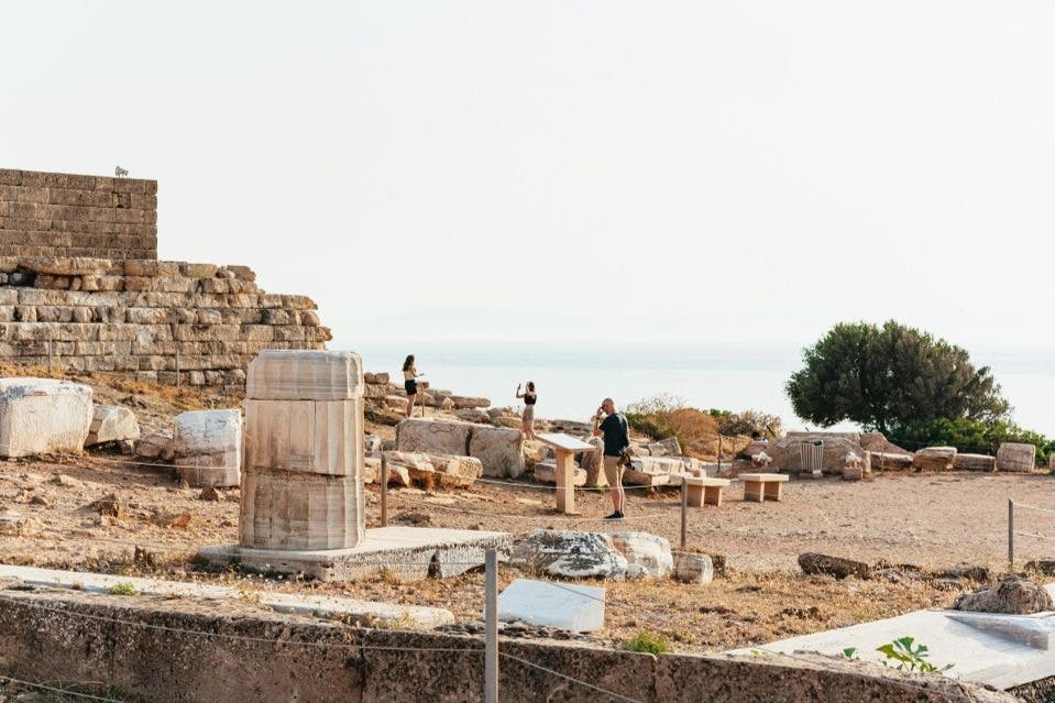 Τουρίστες εξερευνούν αρχαία ερείπια με διάσπαρτες πέτρινες κατασκευές και θέα στη θάλασσα στο βάθος.