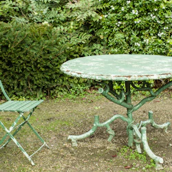 En gammal grön metallstol och ett runt bord i en trädgård, omgiven av täta gröna blad.