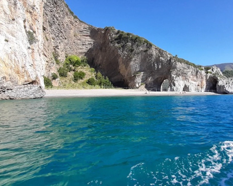 Clear blue waters in the foreground with rocky cliffs and a beach in the background under a clear blue sky.