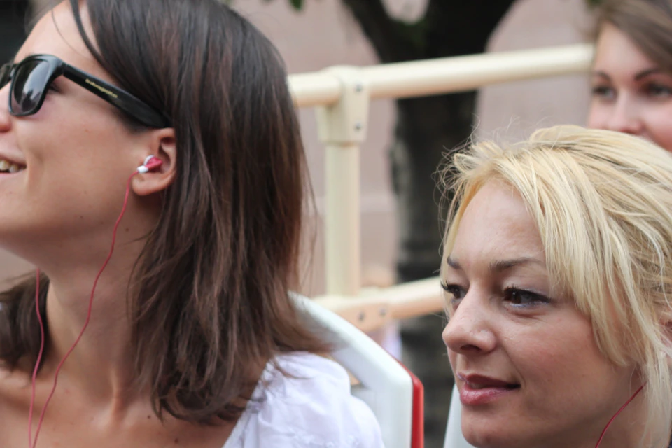 Two women sitting on an outdoor bus wearing earphones, with trees and a building visible in the background.