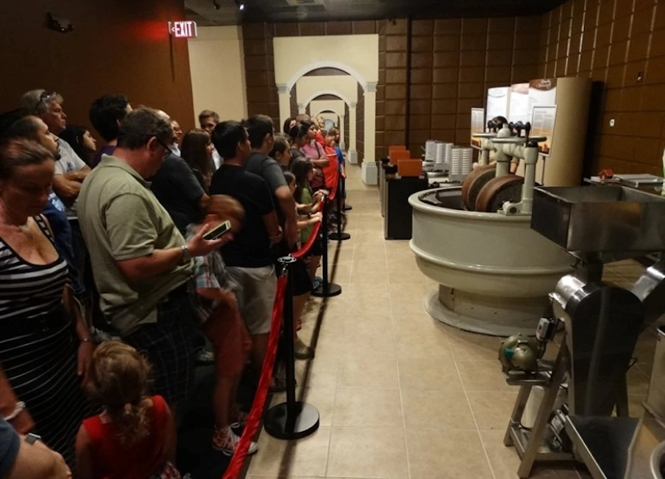 People standing in line inside an exhibit room, observing a large, round industrial machine with informational displays nearby.