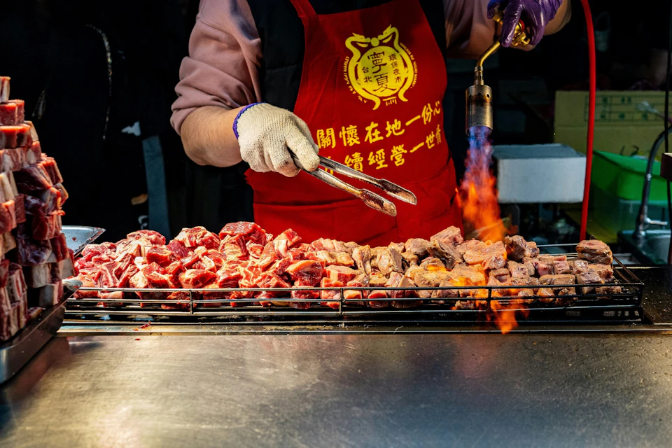 A person torch-grills skewered meat wearing red apron with yellow text, using tongs and a blowtorch.