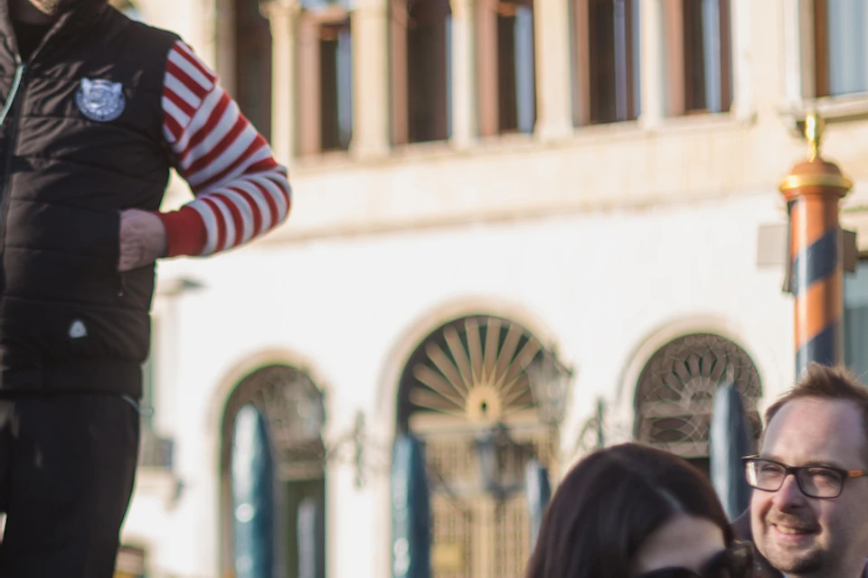 A gondolier stands in a gondola with a couple seated, smiling, and enjoying a scenic ride near historic buildings.