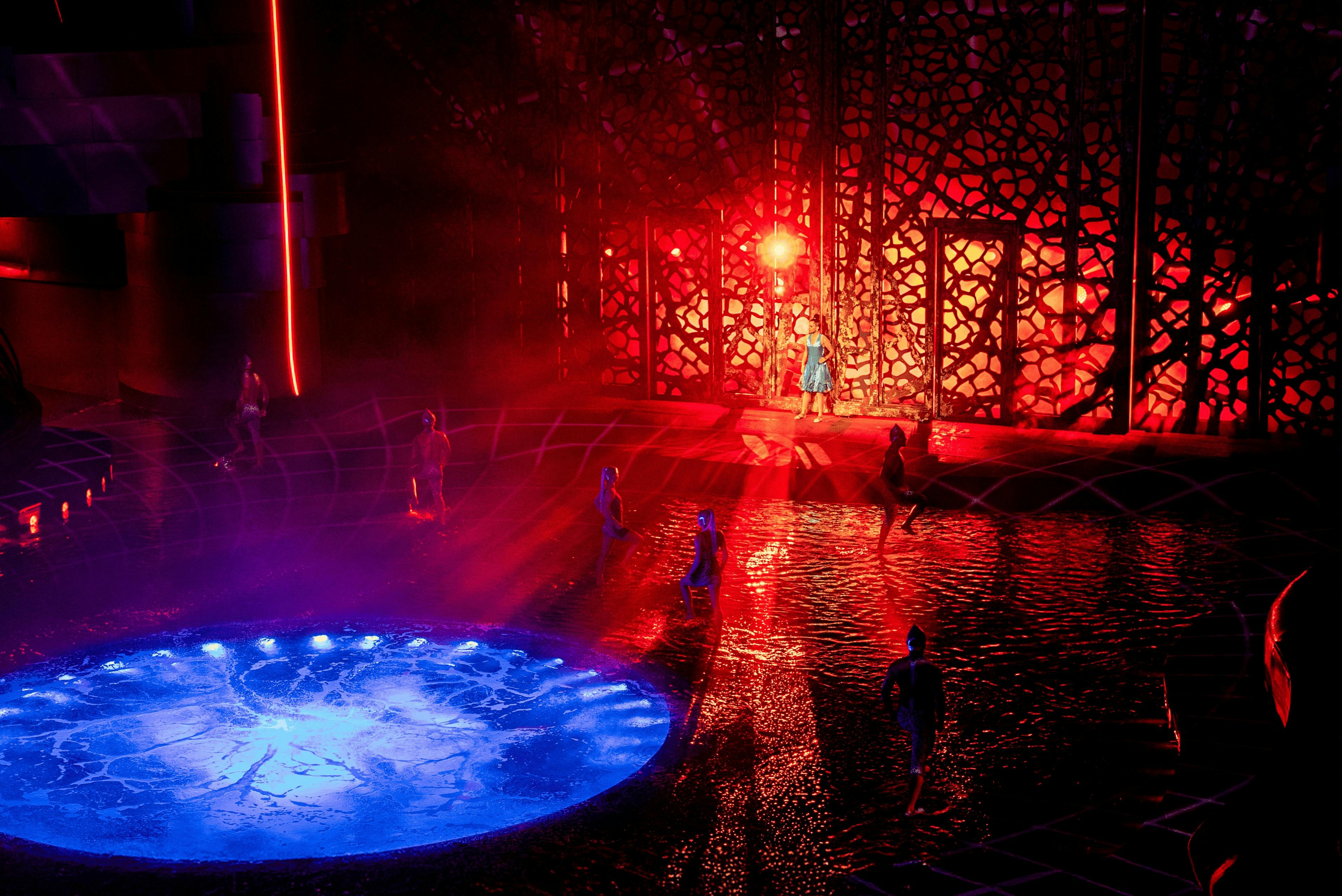 Darsteller in einer Wasserbühnenshow mit dramatischer Beleuchtung, mit einem leuchtend blauen Pool und einem rot beleuchteten Hintergrund.