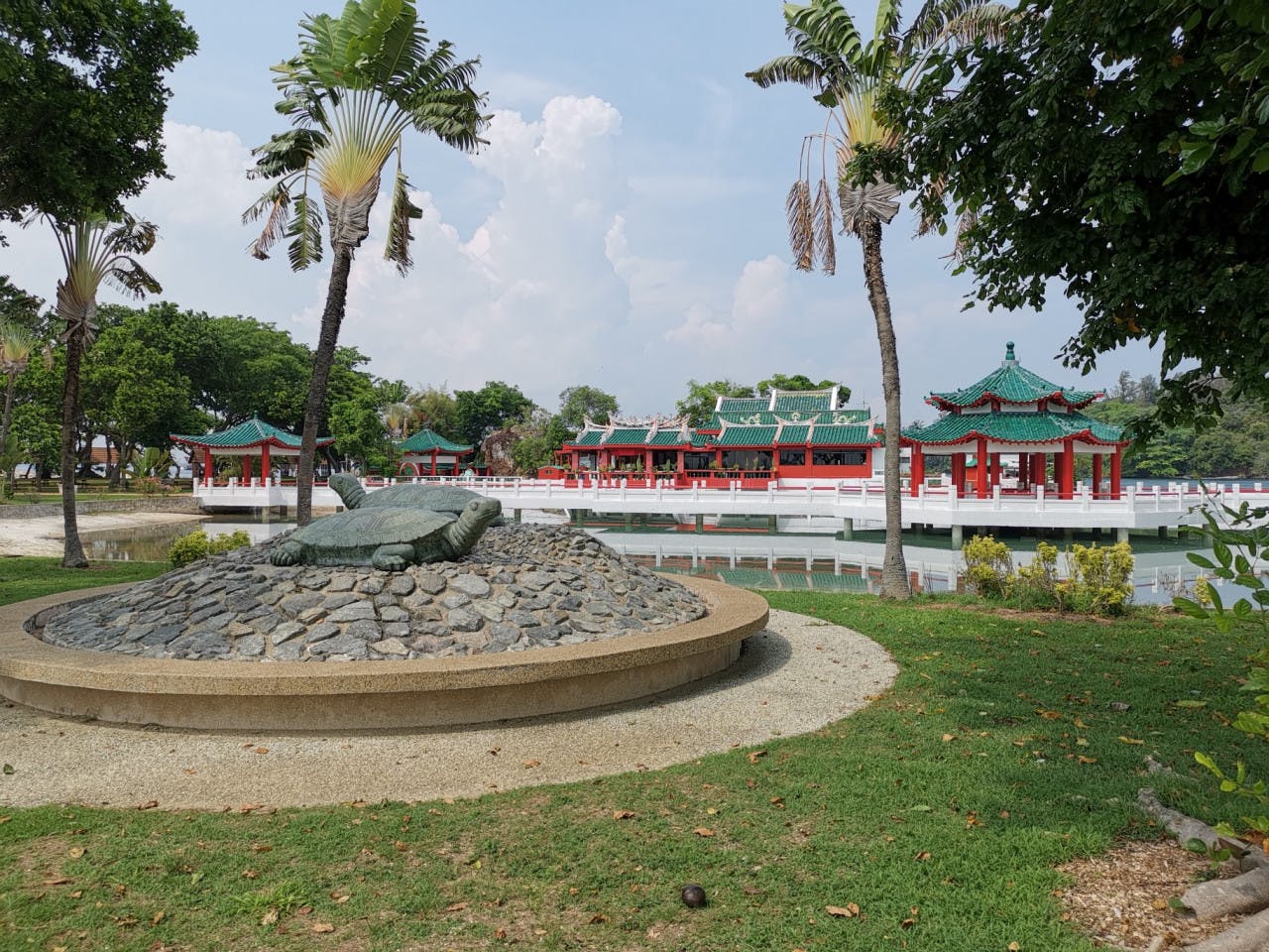 Scéna v parku se sochou želvy na kamenné plošině, palmami, travnatou plochou a tradičními budovami s červenou a zelenou střechou přes rybník.