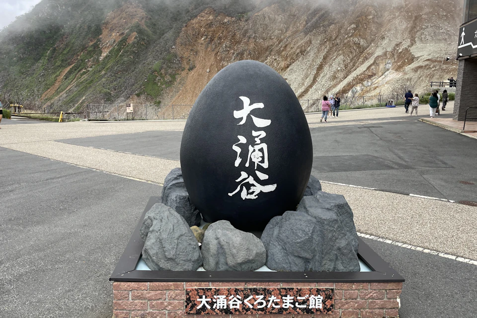 Large black egg-shaped sculpture with Japanese text, surrounded by rocks, with a mountain and people in the background.