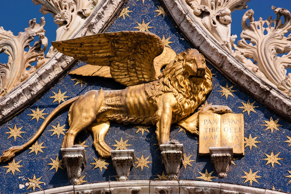 Gold-winged lion sculpture against a blue starry backdrop on an ornate building facade, holding a book with Latin text.