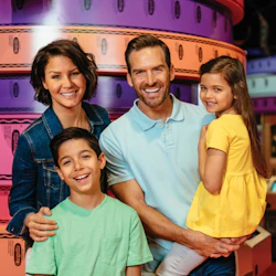 A family of four smiling in front of colorful rolls of paper, with a boy and girl standing with their parents.