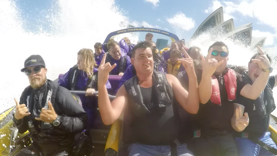 People on a boat ride wearing life jackets, with water splashing around them, some raising their hands and making gestures.