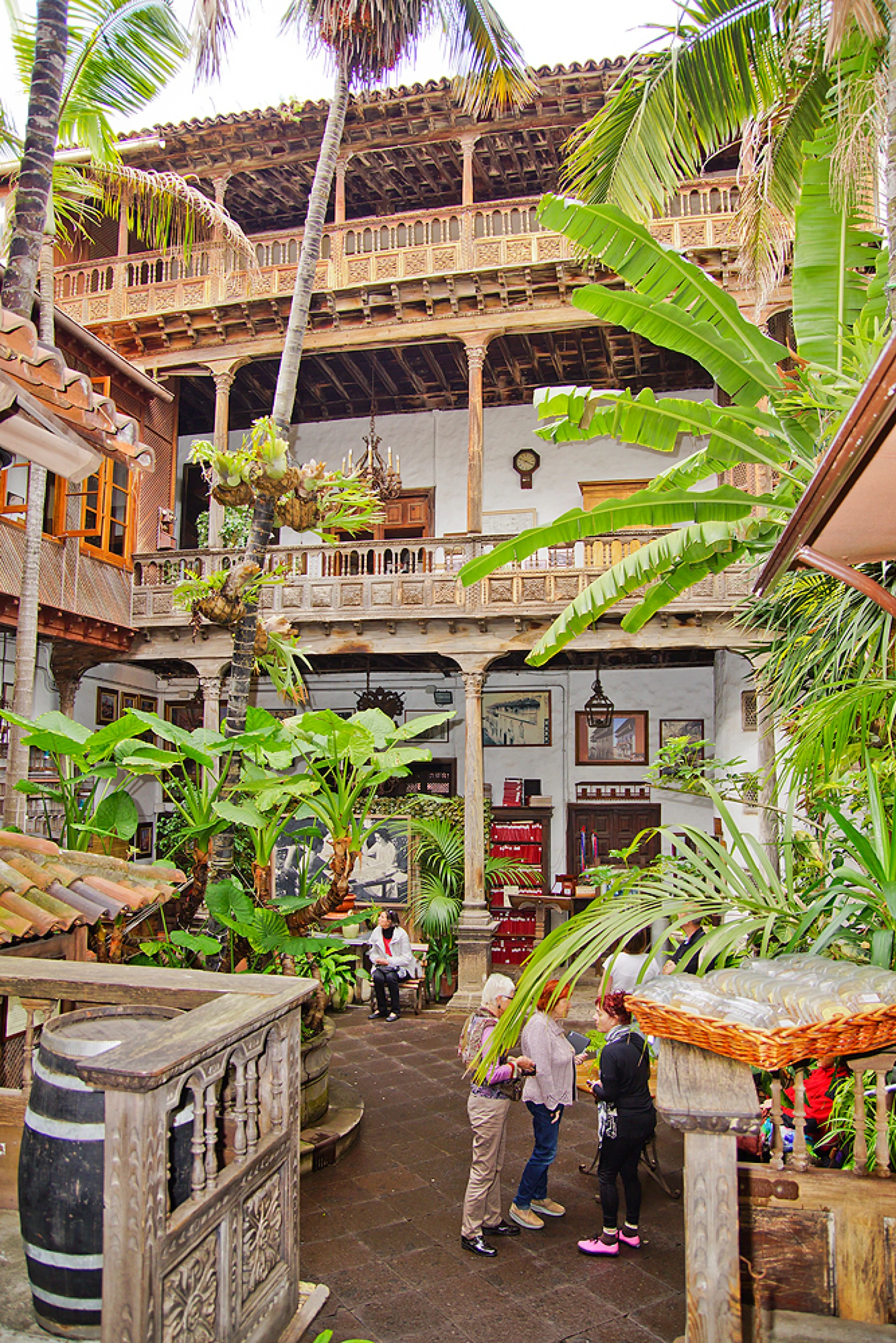 Lush courtyard with people, surrounded by plants and multi-level wooden balconies featuring detailed architecture.