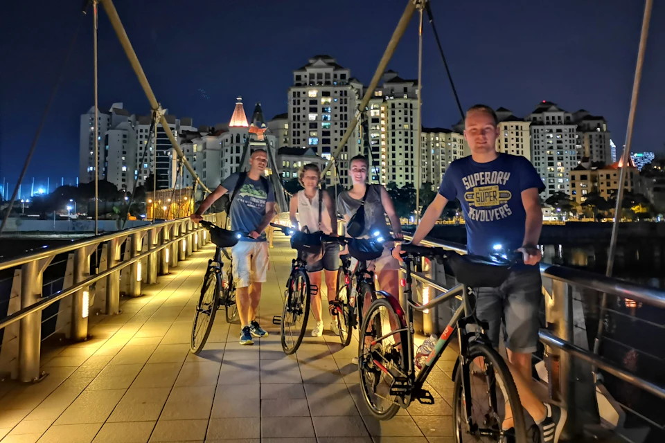 Four people with bicycles on a bridge at night, with city buildings illuminated in the background.