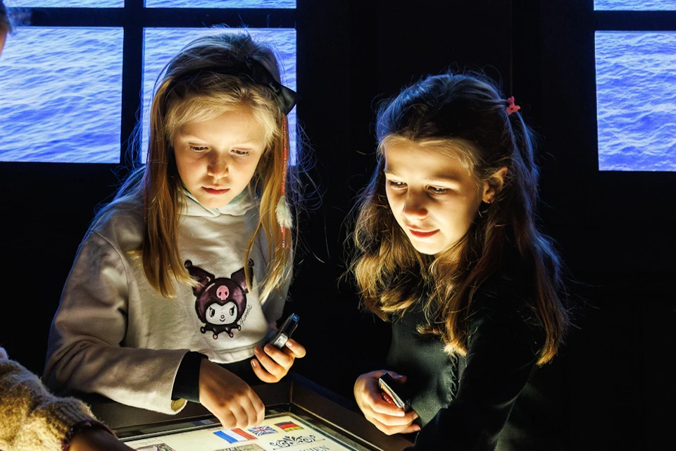 Four children examining a historical map on an illuminated table in a room with windows showing a sea view.