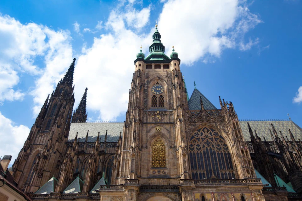 A historic cathedral with ornate architecture, spires, and a large stained glass window under a bright, partly cloudy sky.