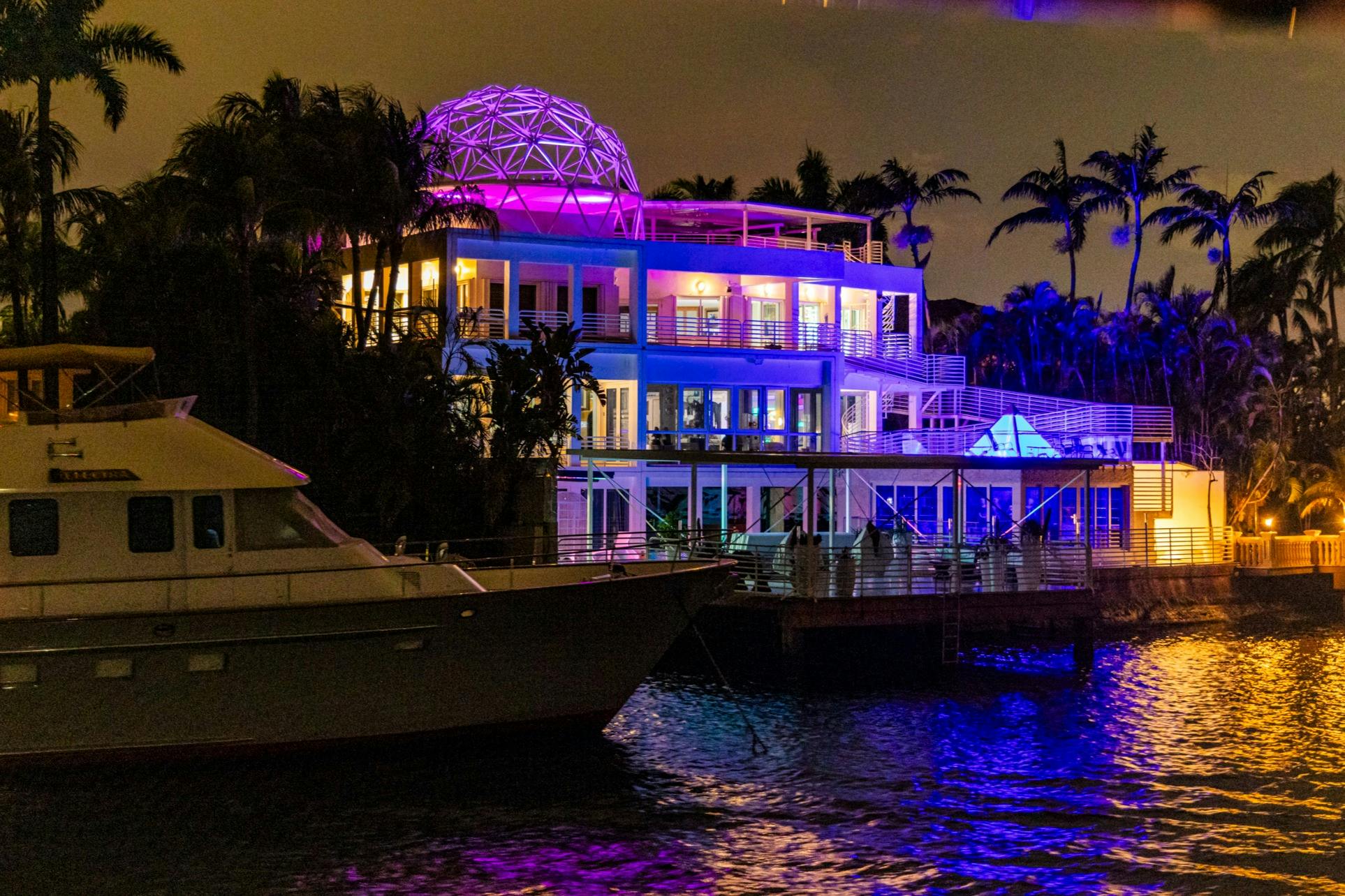 A brightly lit modern building by the water at night, with colorful lights and a geodesic dome on the roof.