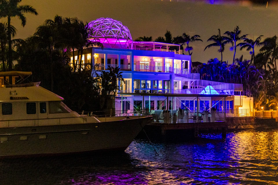 A brightly lit modern building by the water at night, with colorful lights and a geodesic dome on the roof.