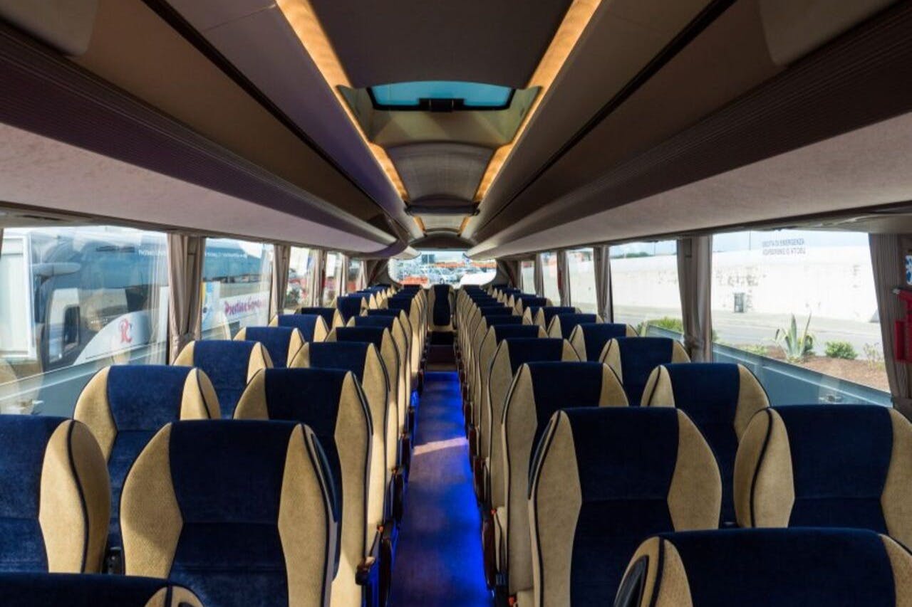 Interior of an empty tour bus with rows of blue and beige seats, overhead lighting, and large windows on either side.