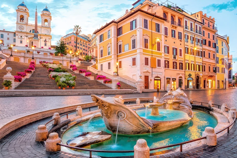 A scenic urban square with a decorative fountain in the foreground and colourful buildings and stairways in the background.