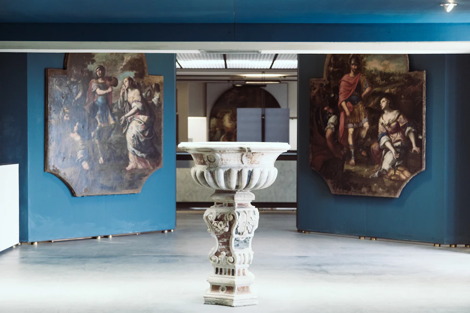 Ornate stone pedestal stands in gallery, flanked by large classical paintings on blue walls.