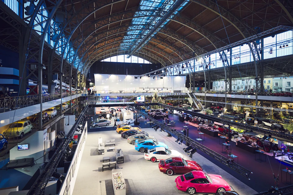 An exhibition hall with sports cars on display, featuring bright lights, high arched ceilings, and various exhibits on the floor.