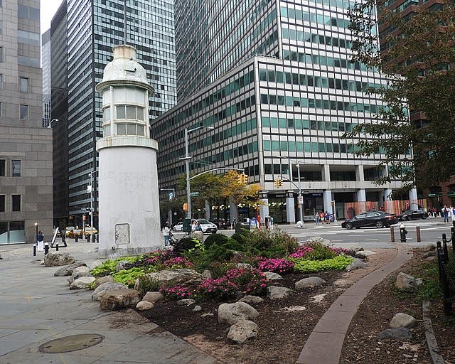 Un petit phare blanc est entouré de parterres de fleurs dans un parc urbain, avec de grands bâtiments et une rue en arrière-plan.