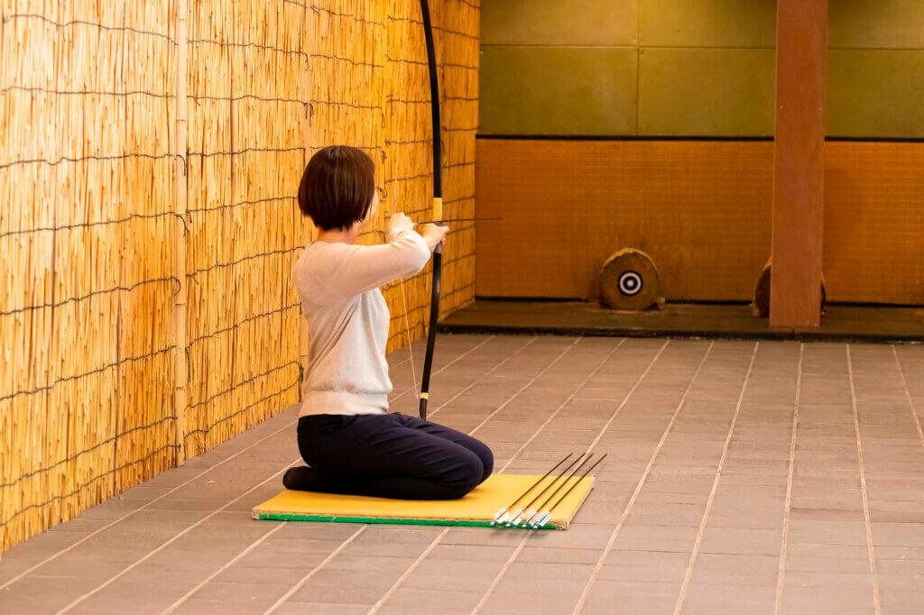 A person kneeling on a mat indoors, holding a bow aimed at a target in a bamboo-paneled archery range.