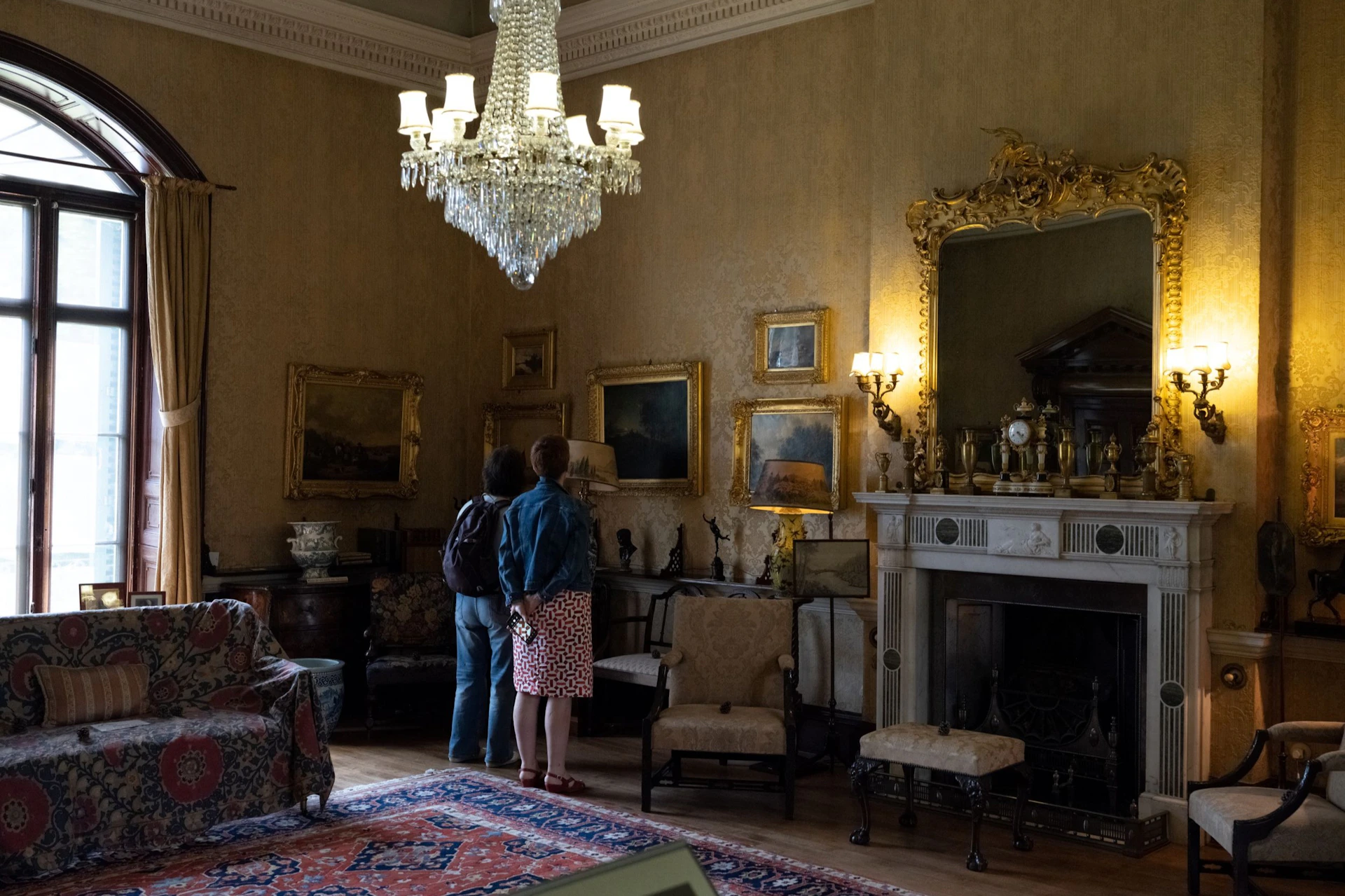 Two people in a vintage room with antique furniture, paintings on the walls, a large chandelier, and an ornate fireplace.