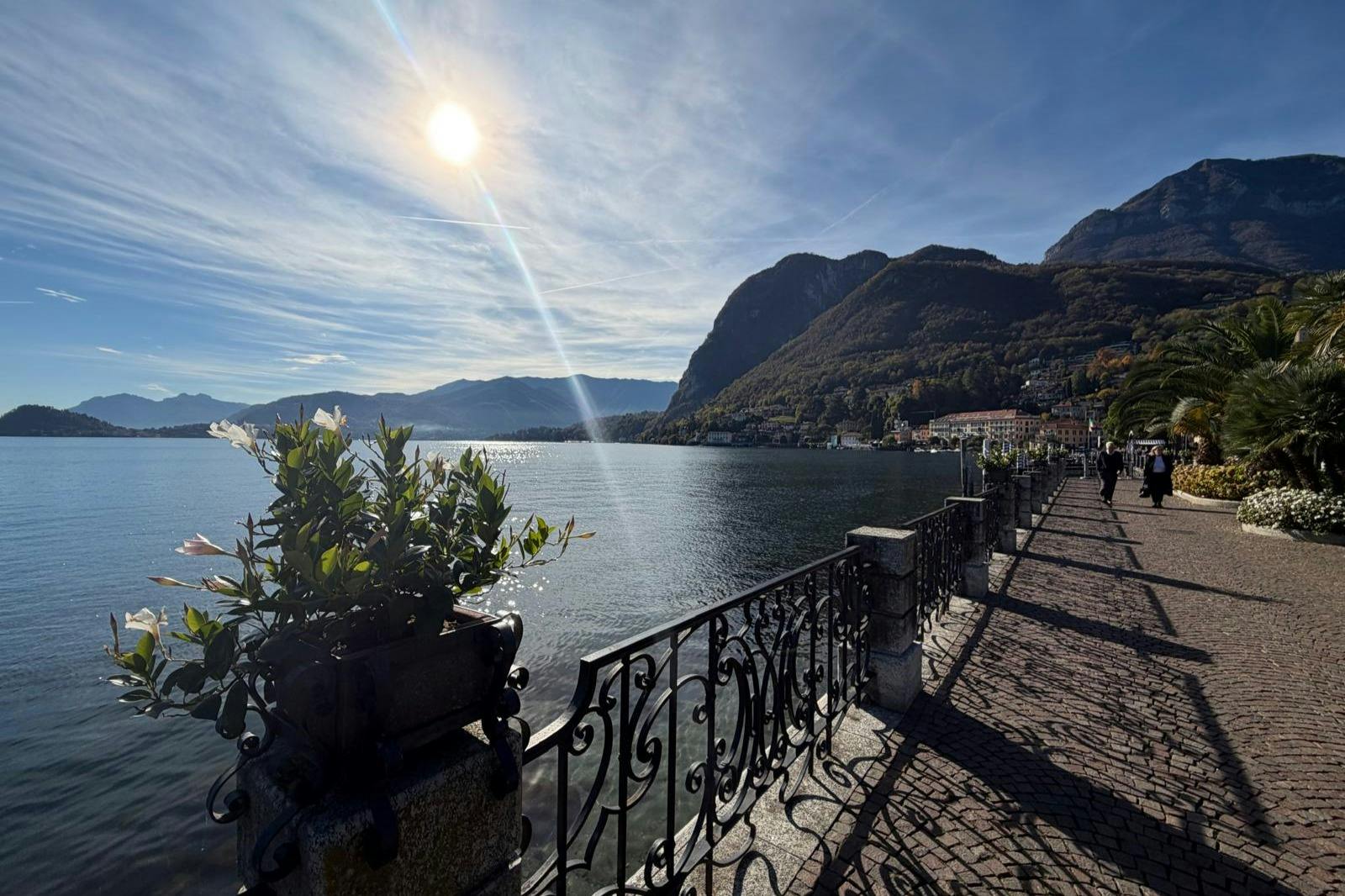 Sunlit lakeside promenade with decorative railing, planters, and distant mountains under a partly cloudy sky. Few people walk along.