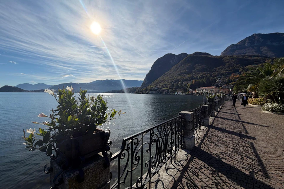 A sunlit lakeside promenade with decorative railings, planters and distant mountains beneath a partly cloudy sky. A few people are walking along it.