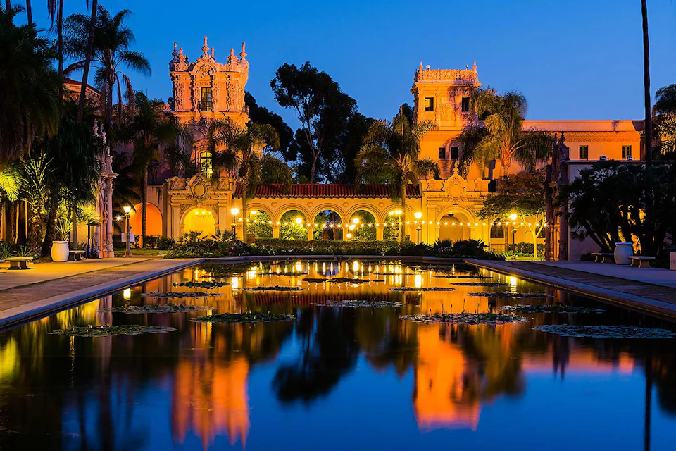 Historic buildings and palm trees with bright lighting reflecting in a tranquil pond during twilight.