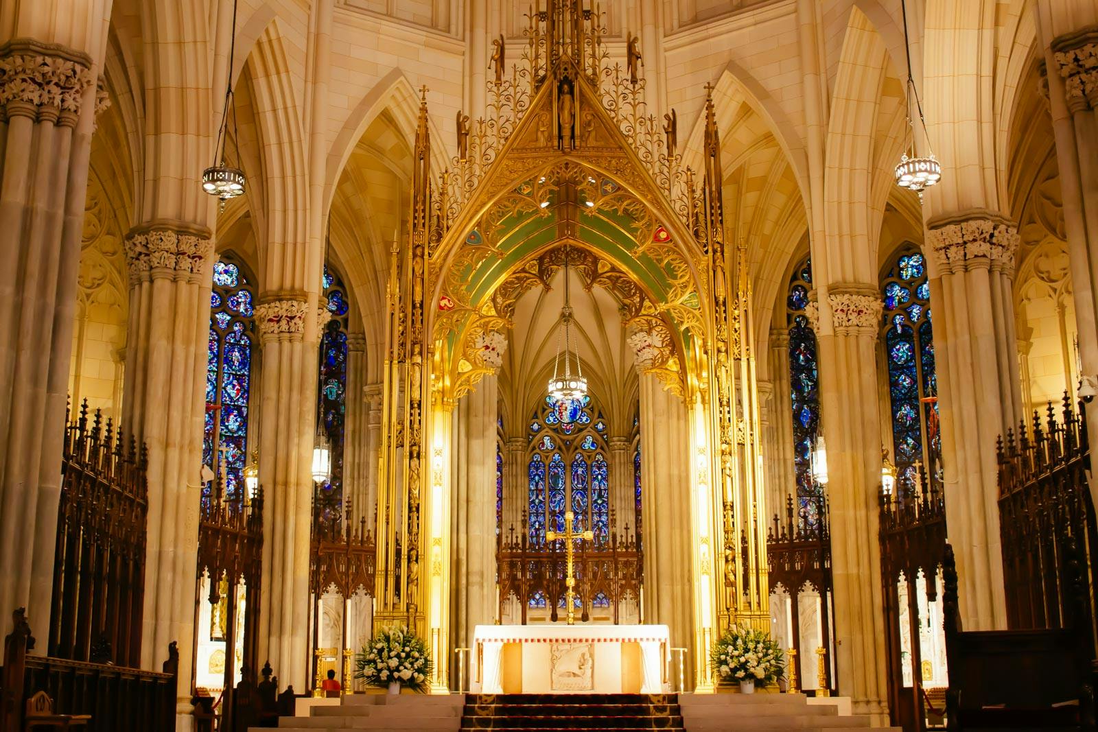 Intricately decorated cathedral altar with golden accents, ornate columns, stained glass windows, and floral arrangements.
