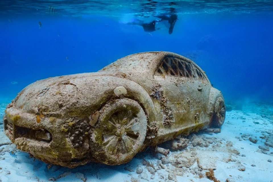 An underwater scene showing a seaweed-covered, submerged car with a scuba diver swimming above in clear blue water.