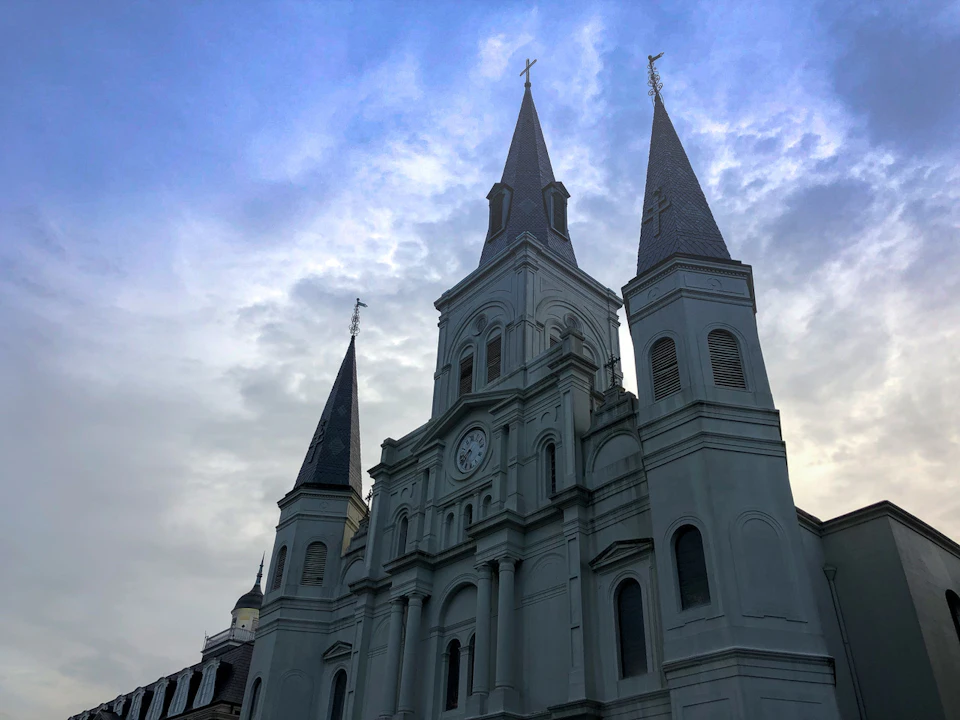 Tall church with three spires, clock on central spire, and a cloudy sky backdrop. Architecture features arched windows.